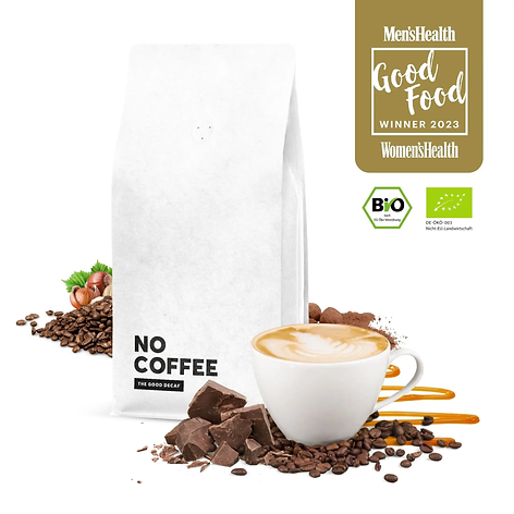 Das Bild zeigt eine stilvolle Präsentation eines Produkts namens "No Coffee". Im Mittelpunkt steht eine weiße Verpackungstüte mit dem Aufdruck "NO COFFEE - The Good Decaf". Die Verpackung ist schlicht gestaltet und vermittelt einen minimalistischen, modernen Eindruck.  Vor der Verpackung liegt eine Tasse Cappuccino mit einem schönen Milchschäumen-Muster, das auf sorgfältige Zubereitung hinweist. Rund um die Tasse und die Verpackung sind Kaffeebohnen, Haselnüsse, Schokoladenstücke und Kakaopulver dekorativ verteilt, was auf die geschmacklichen Noten des Kaffees hindeutet – möglicherweise Haselnuss und Schokolade.  In der oberen rechten Ecke des Bildes befinden sich mehrere Logos und Auszeichnungen, darunter:  Ein "Men's Health Good Food Winner 2023" Siegel, was darauf hinweist, dass das Produkt von Men's Health und Women's Health als besonders gut ausgezeichnet wurde. Ein Bio-Zertifikat mit dem Hinweis auf ökologische Landwirtschaft, was die Qualität und Nachhaltigkeit des Produkts unterstreicht. Das Bild insgesamt vermittelt den Eindruck eines hochwertigen, geschmackvollen und nachhaltig produzierten entkoffeinierten Kaffees.